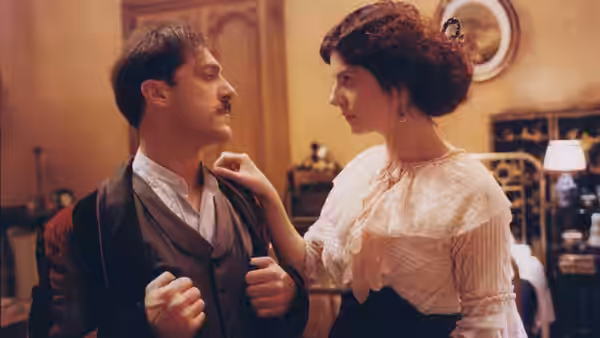 A mustached man adjusts his coat as a woman rests her hand on his shoulder