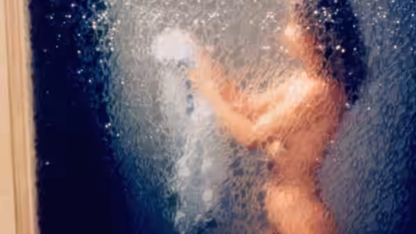 A nude figure moves behind a rain-speckled glass panel