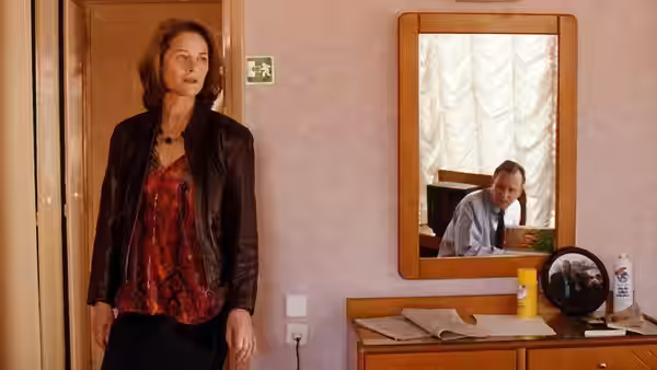 A woman stands beside a dresser as a man appears in the mirror behind her