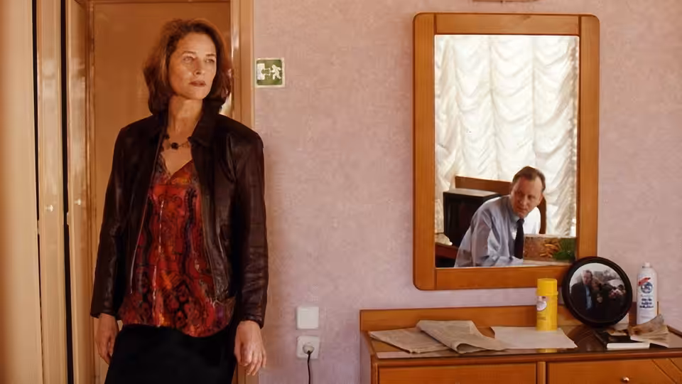 A woman stands beside a dresser as a man appears in the mirror behind her