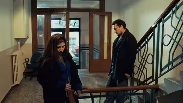 A woman stands by the stairs as a man watches from the lobby