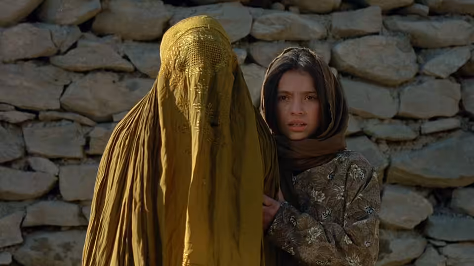 Osama (Marina Golbahari) stands beside a woman in a golden burqa against a stone wall