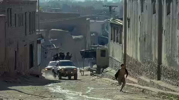 A child runs down a dusty street as vehicles approach between tall buildings