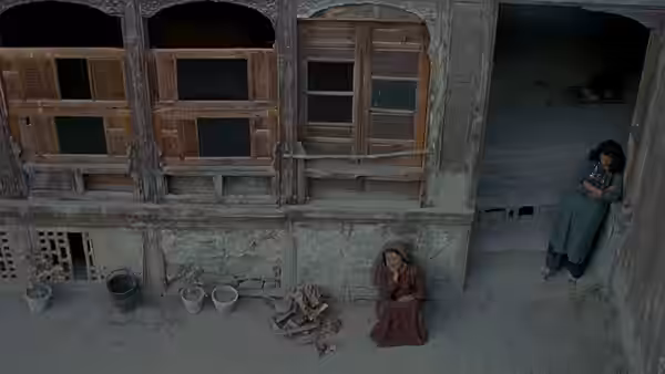 A woman sits beside firewood while another watches from a doorway