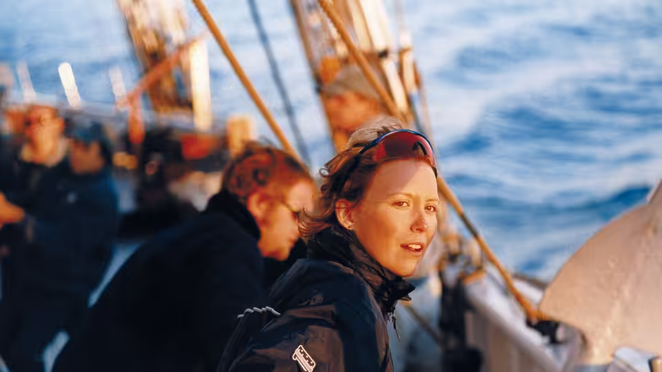 A woman in sailing gear turns toward the camera aboard a boat at sea