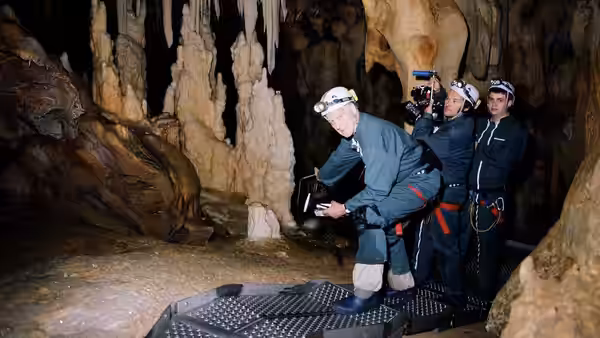 A helmeted cave researcher crouches on a platform as colleagues film inside the cavern