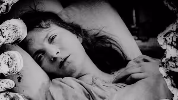 A woman lies among blankets, looking upward as a hand rests beside her