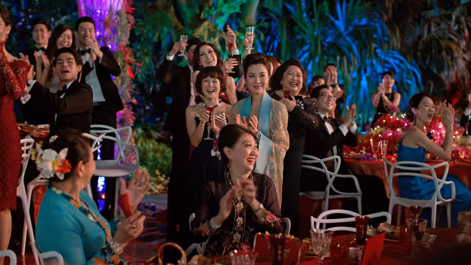 Guests cheer and applaud at an outdoor banquet beneath colorful lights