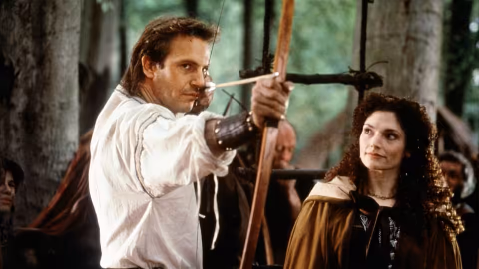 Robin of Locksley (Kevin Costner) aiming a bow with Marian (Mary Elizabeth Mastrantonio) in the woods