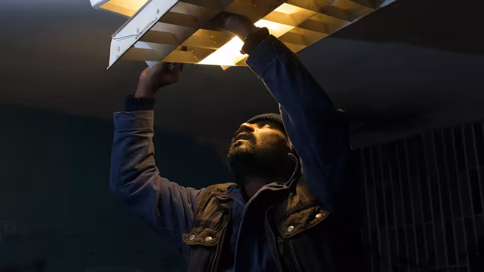 Dheepan (Antonythasan Jesuthasan) reaches up to repair a ceiling light in a dim room