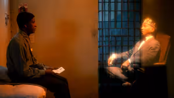 Two men in a prison cell one seated on a bed the other wearing a crown