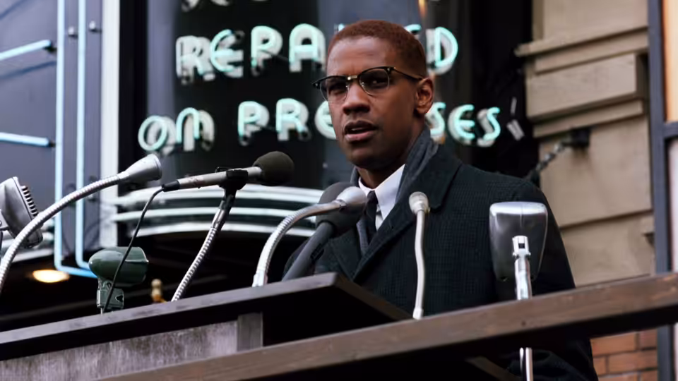 Malcolm X (Denzel Washington) speaking at a podium with multiple microphones