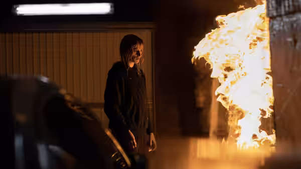 A woman stands beside a wall of flames in a dim garage