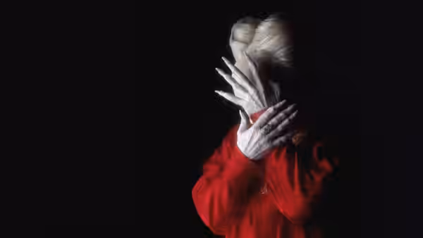 A person in red covers their face with clawed white hands against blackness