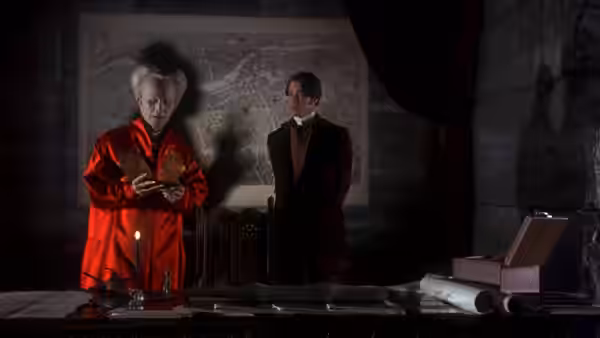 An elderly man in red stands with a suited companion before a large map