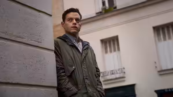 Heller (Rami Malek) leans against a stone wall on a city street