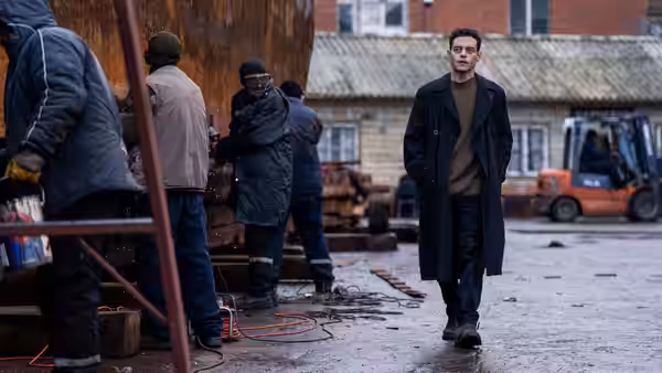 Heller (Rami Malek) walks past workers in an industrial yard