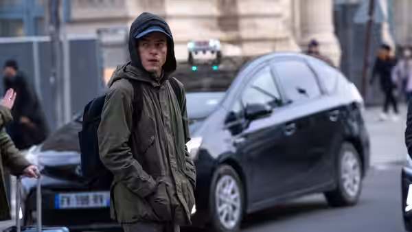 Heller (Rami Malek) in a hooded jacket watches the street beside parked cars