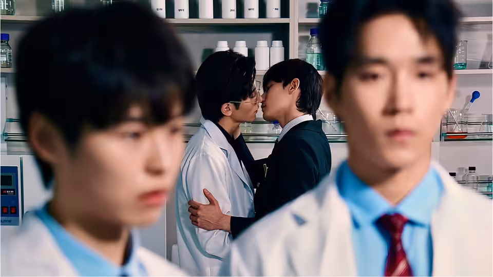 Two students kiss among laboratory shelves, with two classmates blurred in the foreground
