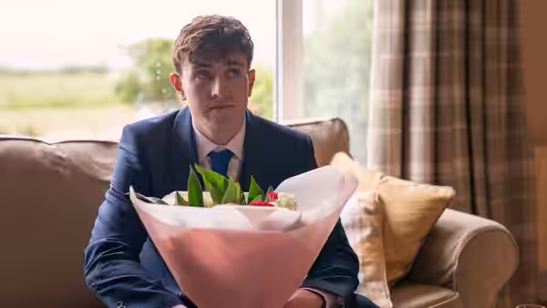 A young man in a suit holds a bouquet while seated on a sofa