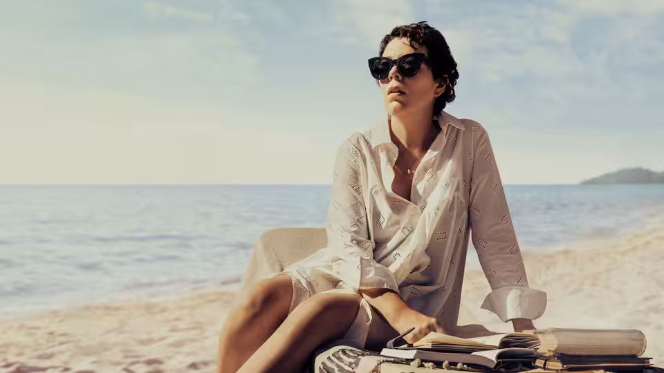 Leda Caruso (Olivia Colman) sits beside her books on the beach, looking out to sea