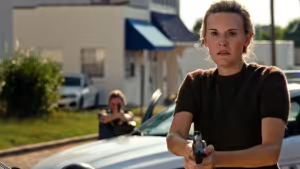 Amelia Pembrock (Maggie Grace) holds a handgun as a man aims behind her