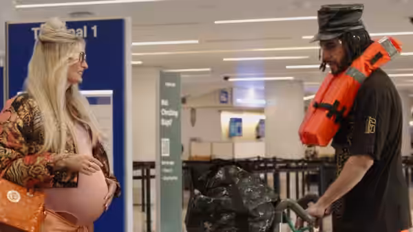 Ali G (Sacha Baron Cohen) pushes a luggage cart beside a pregnant blonde woman at an airport