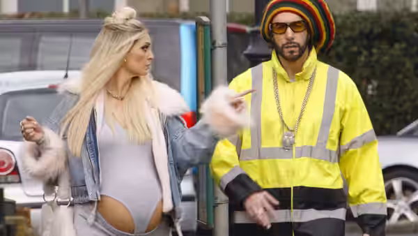 Ali G (Sacha Baron Cohen) walks beside a blonde woman in a high-visibility jacket