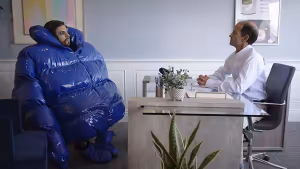 A bearded man in an oversized blue inflatable suit sits across a desk from a doctor