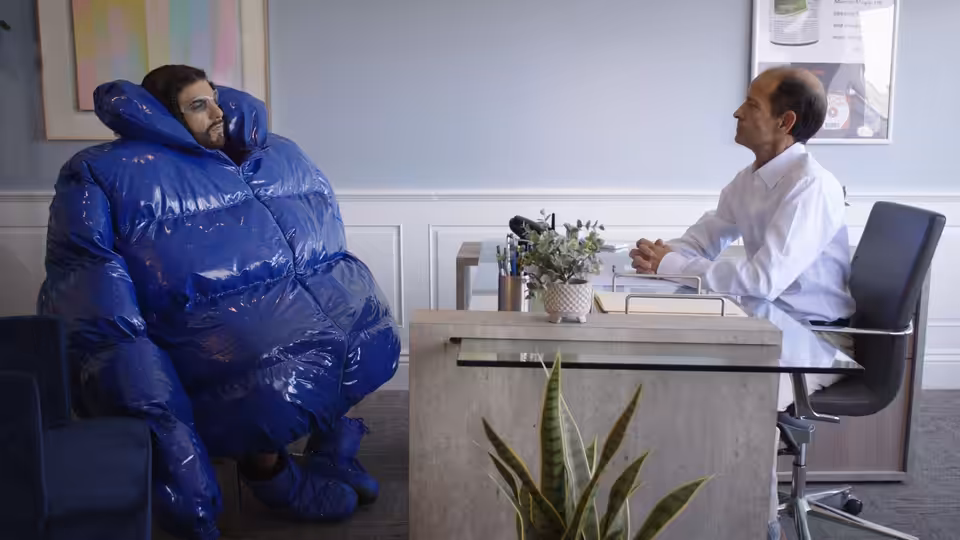 A bearded man in an oversized blue inflatable suit sits across a desk from a doctor