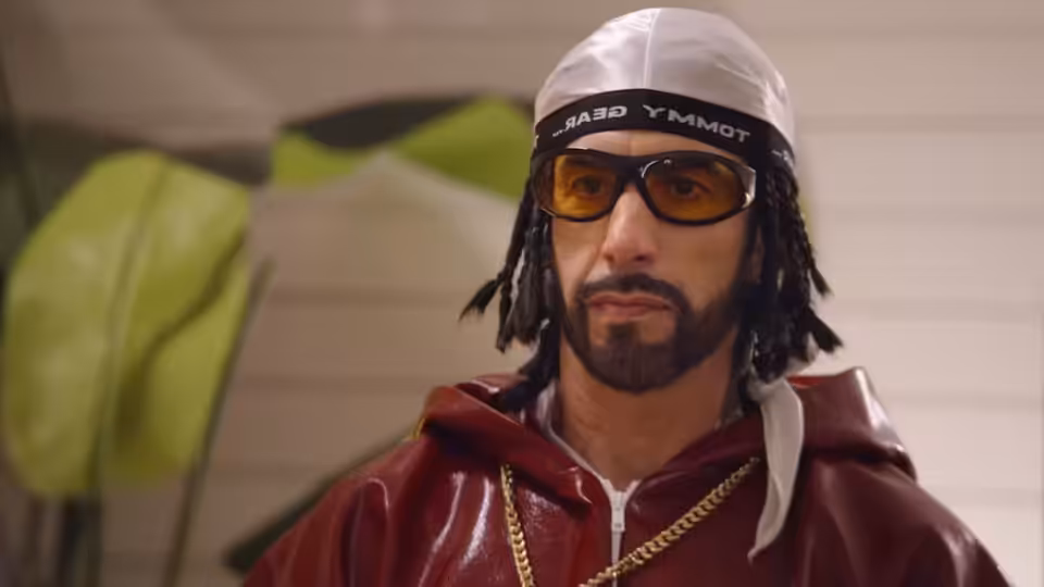 Ali G (Sacha Baron Cohen) wearing a white cap, tinted glasses and gold chains indoors