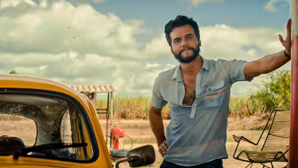Wagner Moura stands by yellow car in rural field under partly cloudy sky