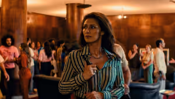 Woman in striped shirt stands in crowded bar with people dancing behind