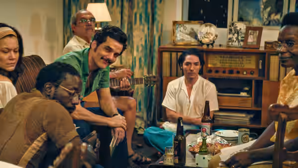 Group sits in living room while man plays guitar among bottles and food