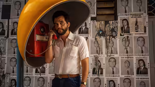 Wagner Moura talks on red telephone inside yellow booth against wall of portraits