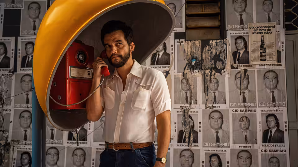 Wagner Moura talks on red telephone inside yellow booth against wall of portraits