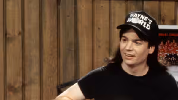 Wayne Campbell (Mike Myers) speaking while seated in front of a wooden wall