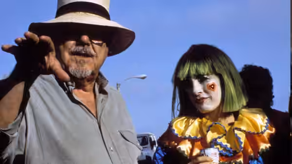 Older man in hat gesturing beside a woman in colorful clown costume