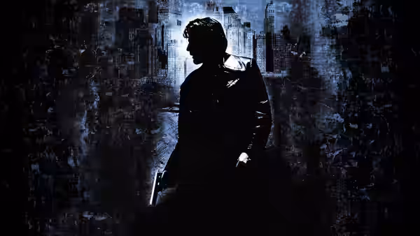 A man in a long coat holds a gun standing against a dark wall