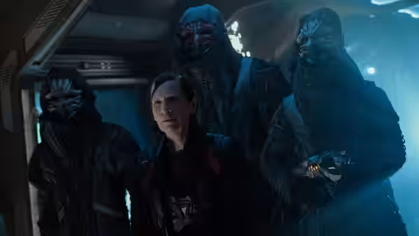 A woman stands surrounded by three masked figures in a dimly lit corridor