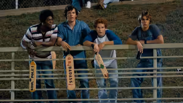 Four young men leaning on a railing holding paddles at a sports field
