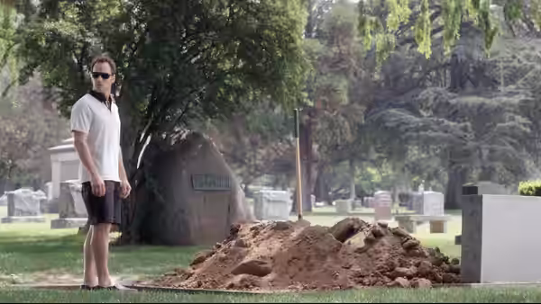A man in sunglasses stands beside a mound of earth in a cemetery