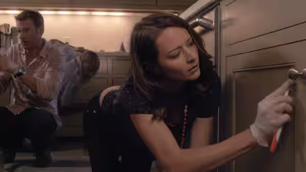 A woman in gloves works a key in a kitchen cabinet while two men crouch behind her