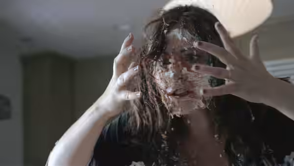 A woman with cake smeared across her face raises both hands in a kitchen