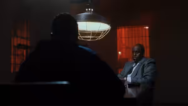 A man in a suit sits at a table facing another man in a dim room