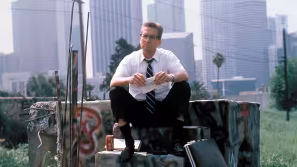 D-Fens (Michael Douglas) seated on concrete rubble holding papers before skyscrapers