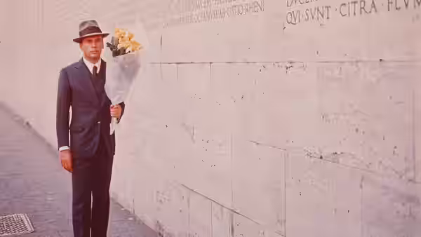 A man in a suit and hat holds flowers beside a stone wall