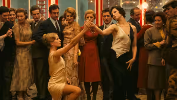 A woman in gold kneels and reaches toward another at a crowded party