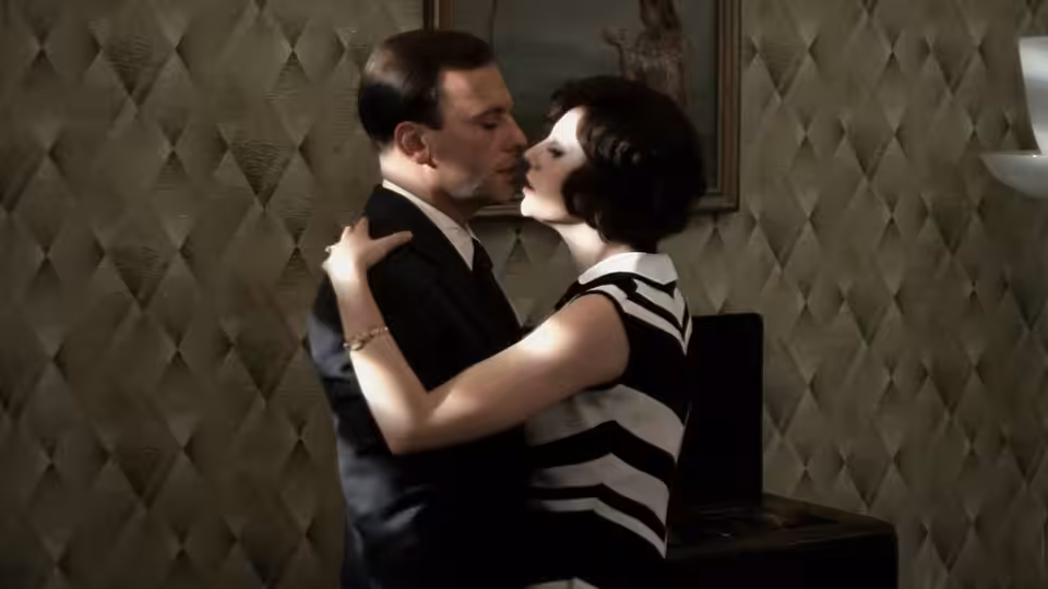 A man in a suit embraces a woman in a striped dress indoors