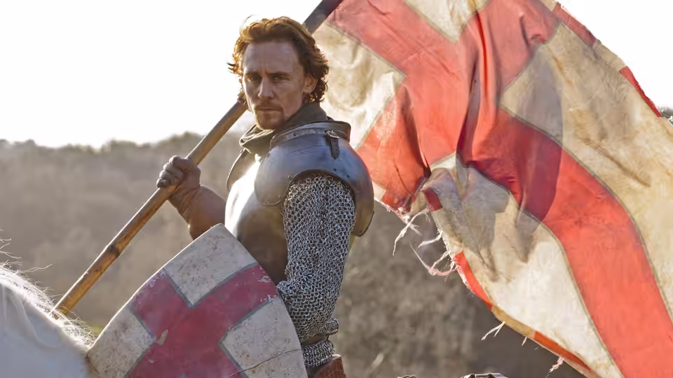 An armored man holds a red-and-white banner in a wooded field